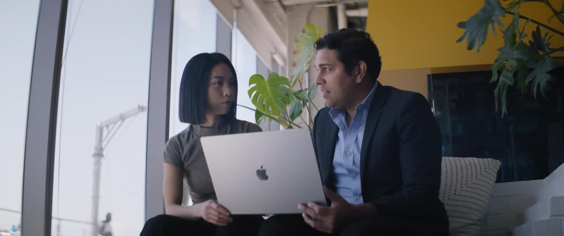 Man and woman sitting side by side in an indoor working environment talking together with a laptop