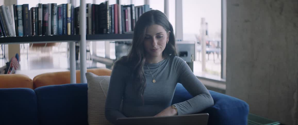 A woman sits on a couch in a library talking to someone via laptop
