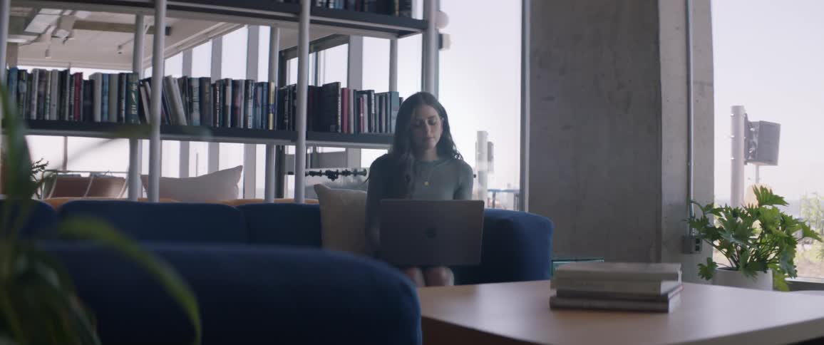 Woman sitting on a blue sofa with books on shelves around her using a laptop