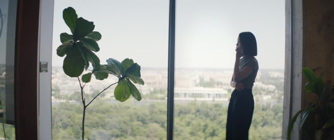 A woman is talking on the phone that is at her ear and she is in front of a large glass window