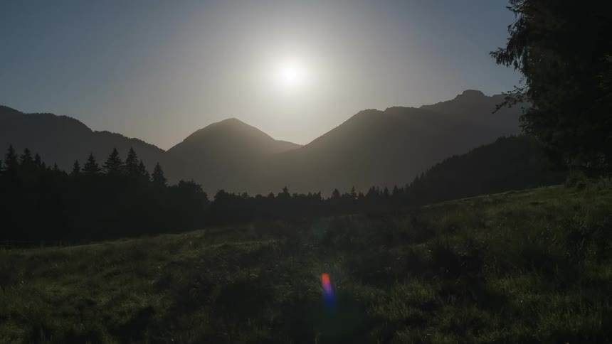 Natural landscape with mountains in the background and a coniferous forest in the foreground under a clear sky with the moving sun