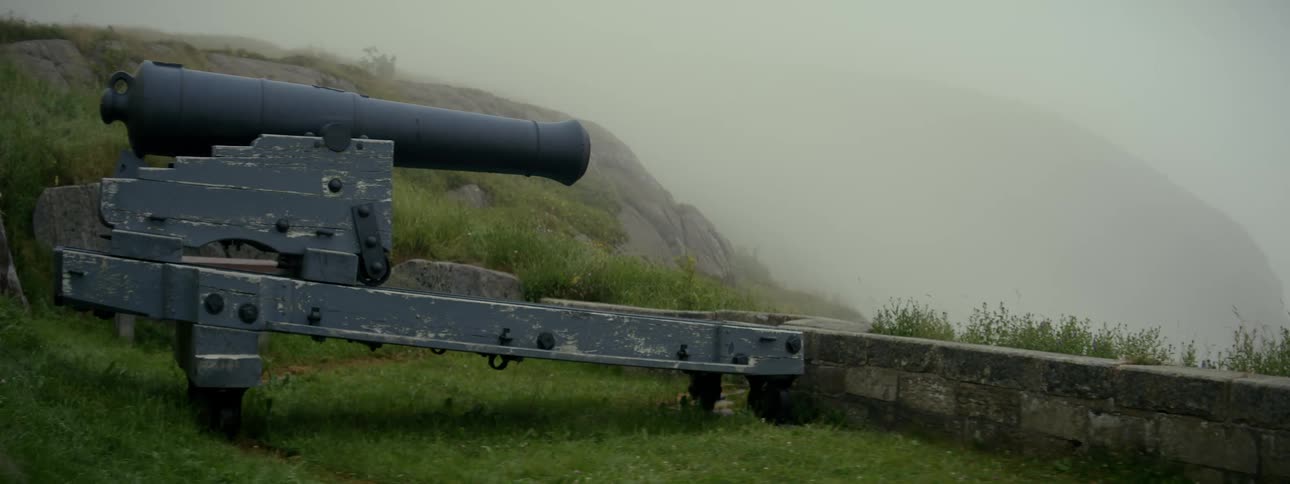 A large cannon and platform resting against a small brick wall on a grassy plain