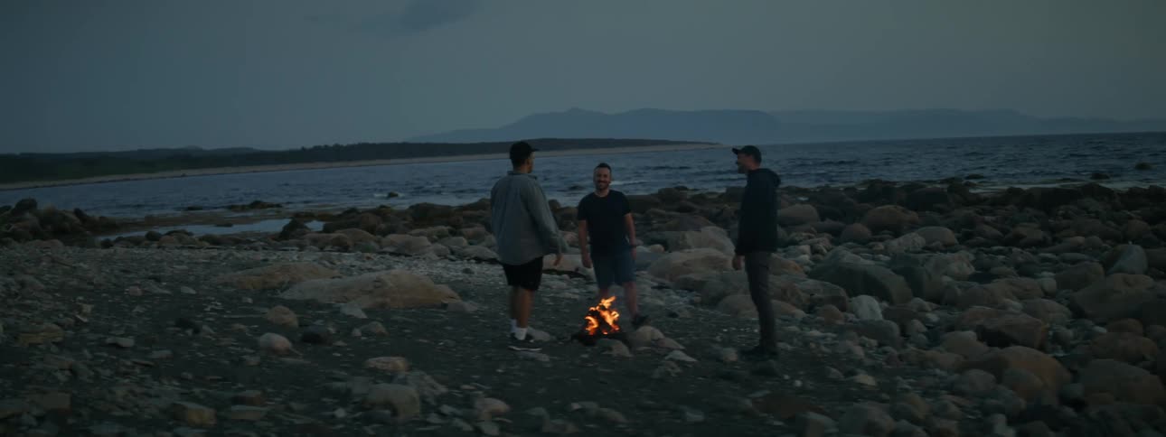 Three men stand on a rocky beach with a campfire between them next to the coast at dusk