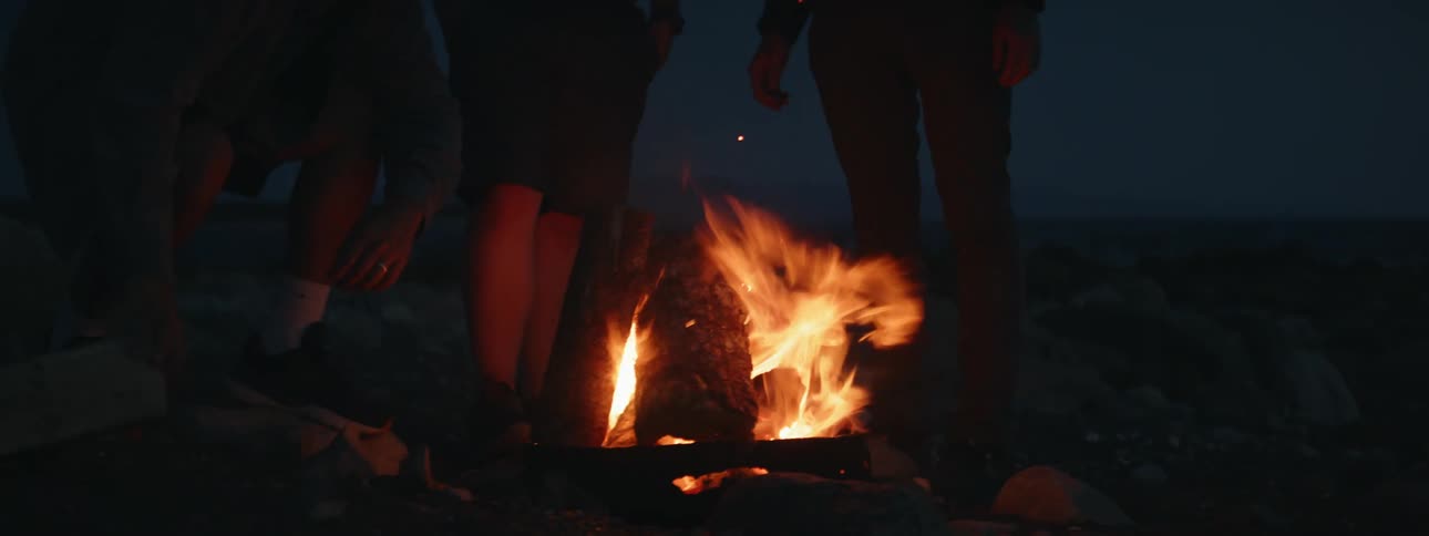Three men are gathered in front of a bonfire on a night in the sand and one of them is adding wood to the fire