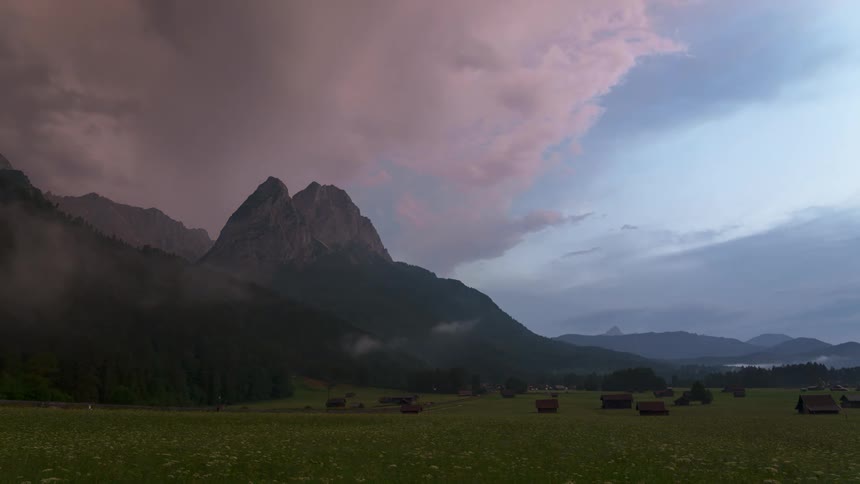 A mountain range under a rapidly moving cloudy sky with small structures dotting a green field