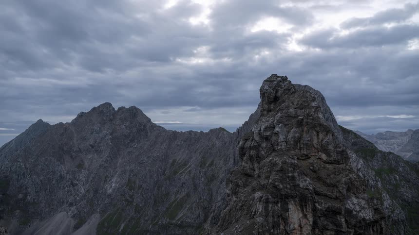 A mountain landscape with a rocky peak in the foreground set against a rapidly moving cloudy sky