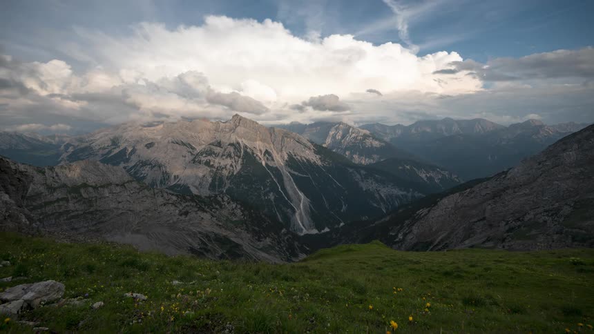 Big white clouds together with think gray ones move across the sky above a mountain range with a meadow in the foreground