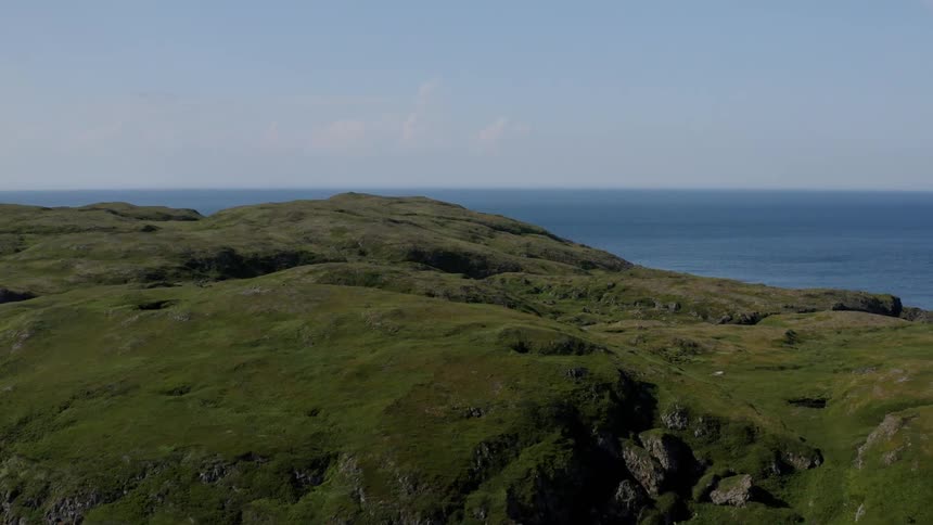 A hill with green grass facing the sea and on the other side there is a calm ocean and many