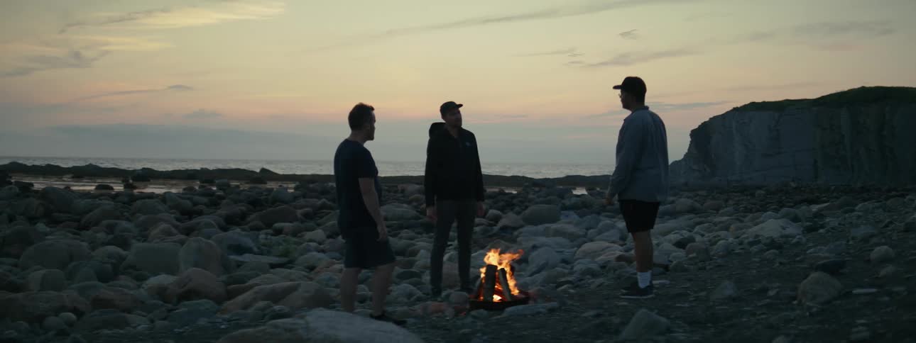 Three men around a campfire surrounded by rocks under a twilight sky