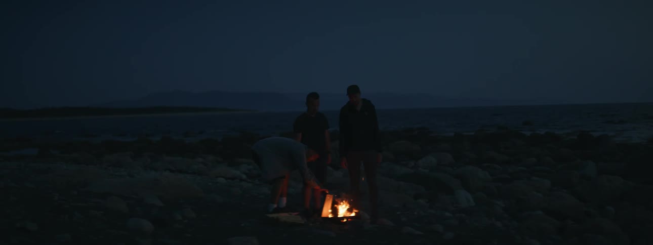 Three men around a campfire in a natural setting by the rocks at night