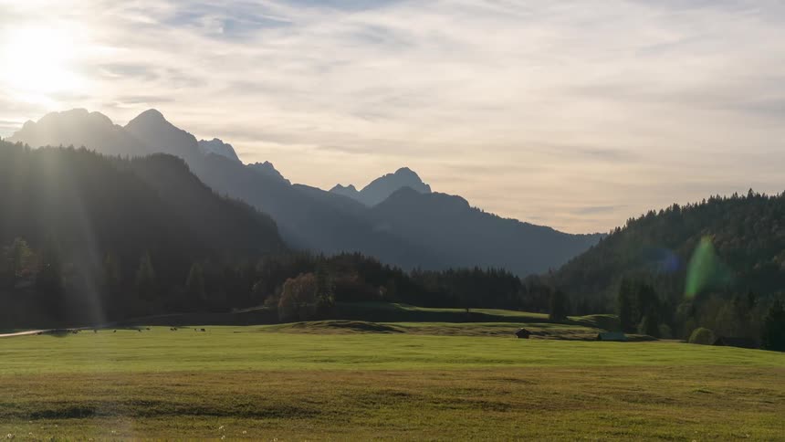 A landscape of a grass field with forests on the sides and a mountain range in the background and clouds passing quickly in the sky