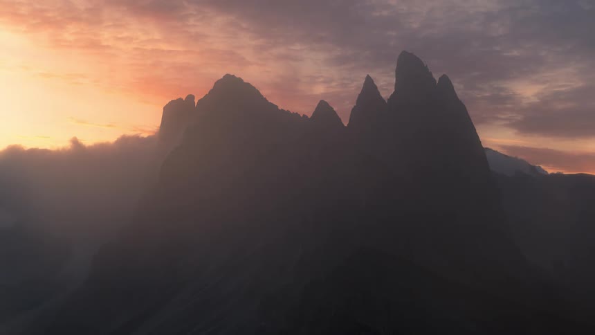 A mountain range with clouds passing by at their peak and the sky with clouds passing quickly and is orange