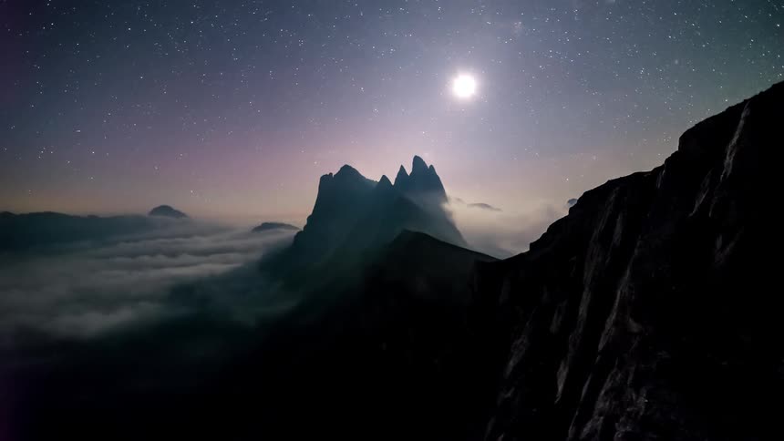 Clouds flow past tall mountains under a bright moon as night turns to dawn