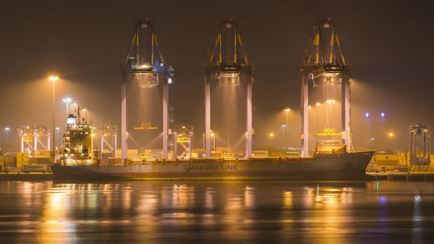 Maersk line cargo ship docked in a night port illuminated by yellow lights with three large container cranes lifting containers
