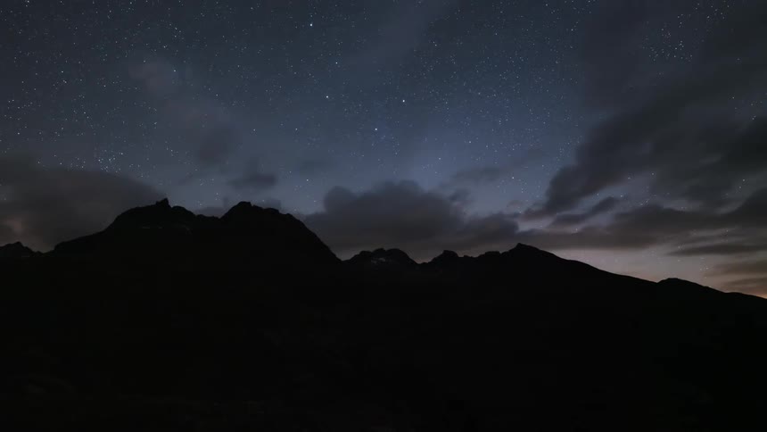 A silhouette of a mountain in the dark and the sky is starry and with several clouds passing by