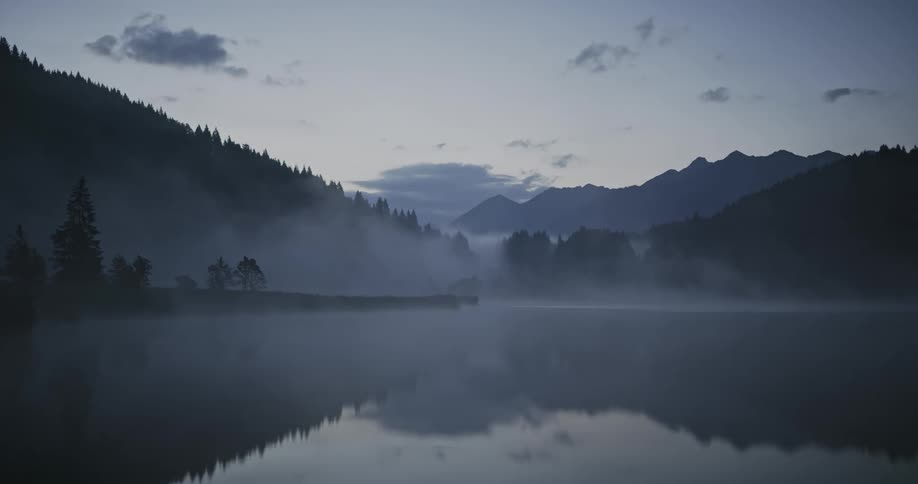 A lake with a little fog above in a region with mountains with trees in the evening