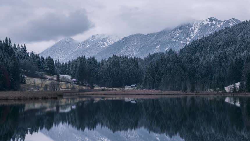 Small cabin stands between forested slopes and reflective lake waters on a foggy winter day