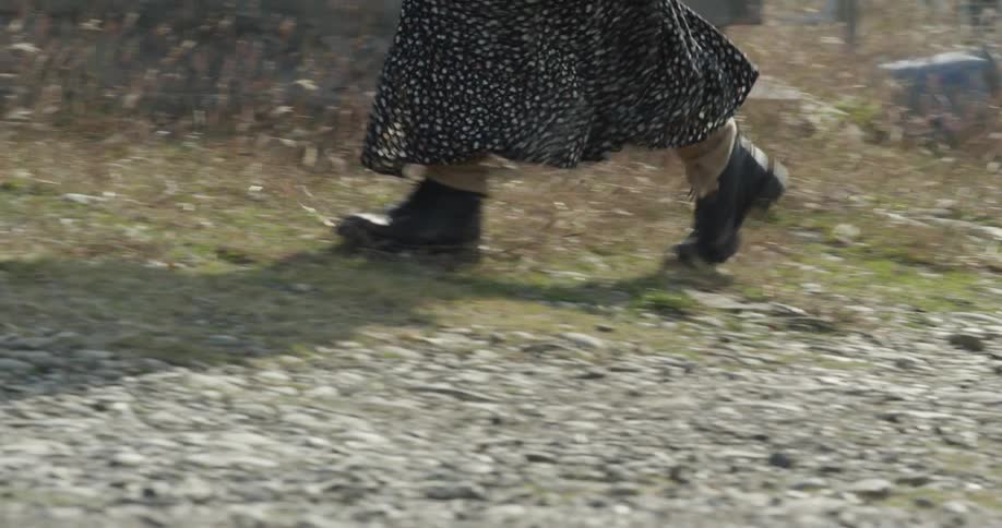 The legs of a person in a skirt walking on a ground of low grass and stones