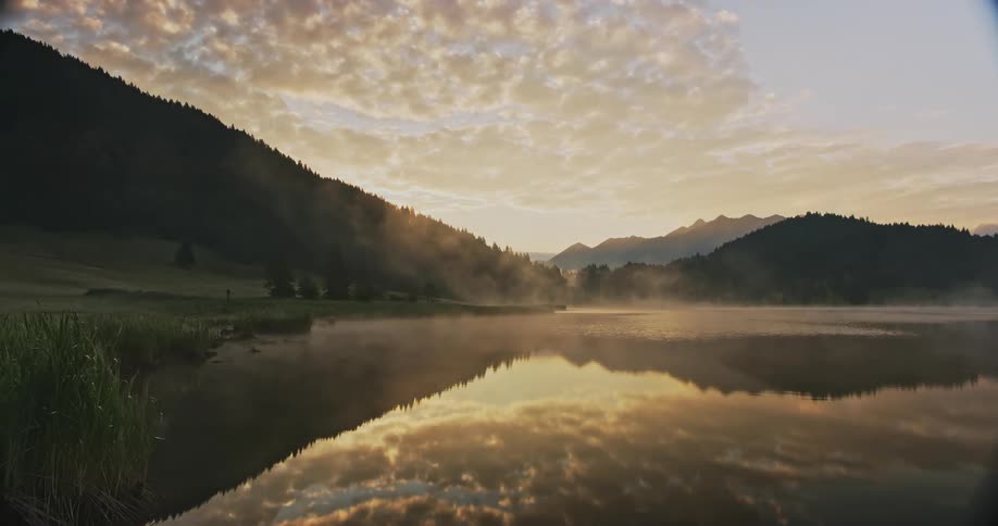 A landscape with misty water in the foreground surrounded by grass and trees mountains rise in the background under a sky with golden clouds at sunrise or sunset