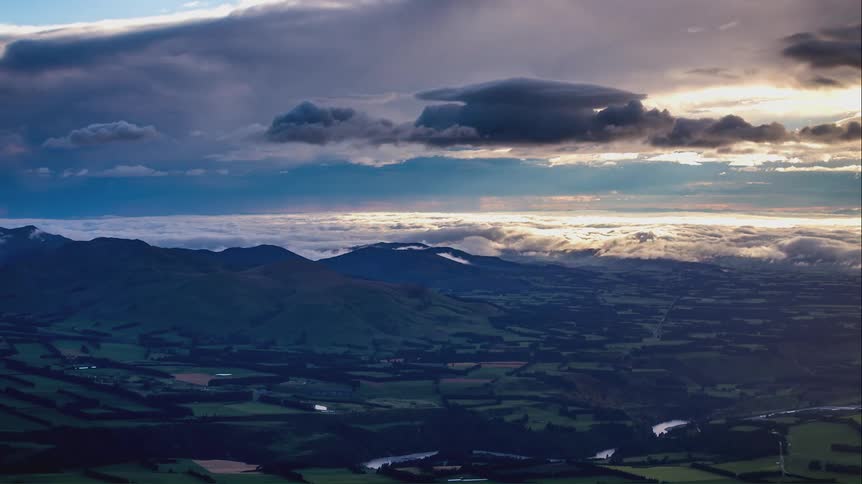 A landscape of several green fields and in the sky there are several white and gray clouds heading towards the right