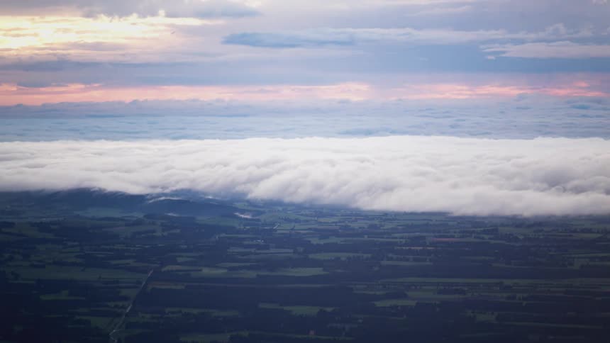Aerial view of thick clouds over green fields with the horizon tinted by the colors of dawn