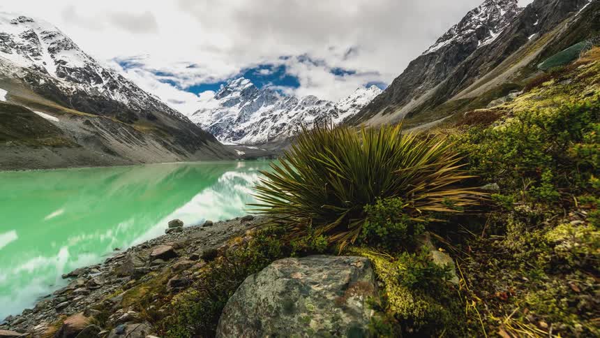 Turquoise green lake surrounded by snowy mountains and low vegetation under a partly cloudy sky