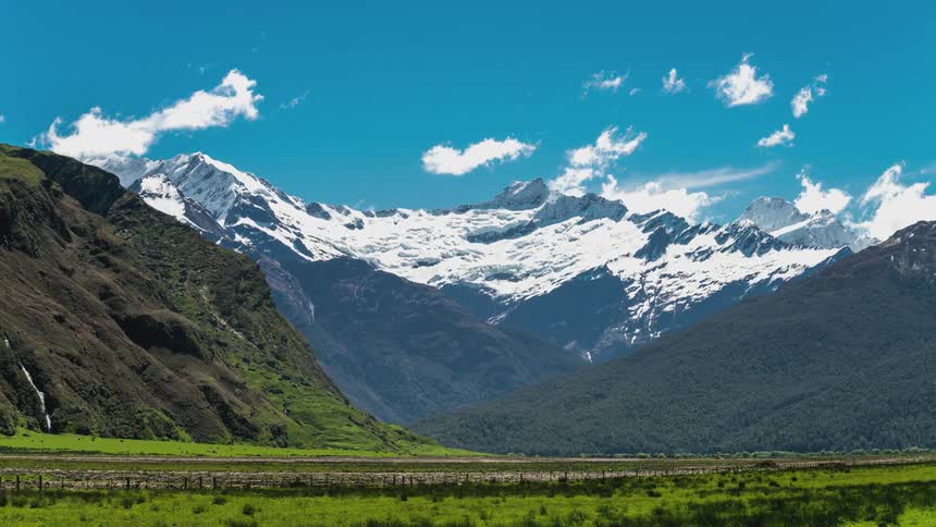 Green valley under snowy mountains under a clear blue sky with scattered clouds