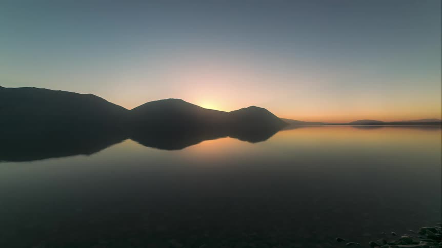 A lake reflecting some mountain silhouettes in the morning and the sun rises behind one of the mountains