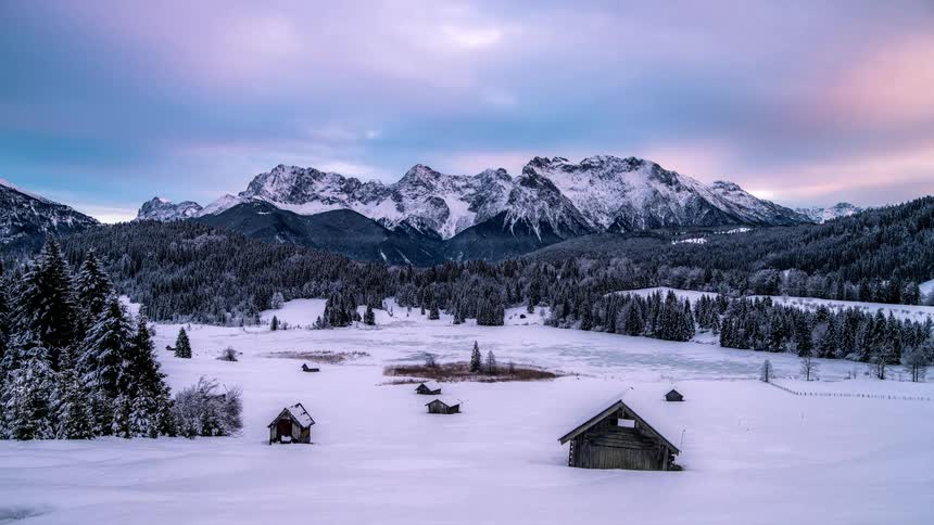 Isolated wooden huts on a snowcovered ground surrounded by conifers and mountains and the sky turning purple at dusk