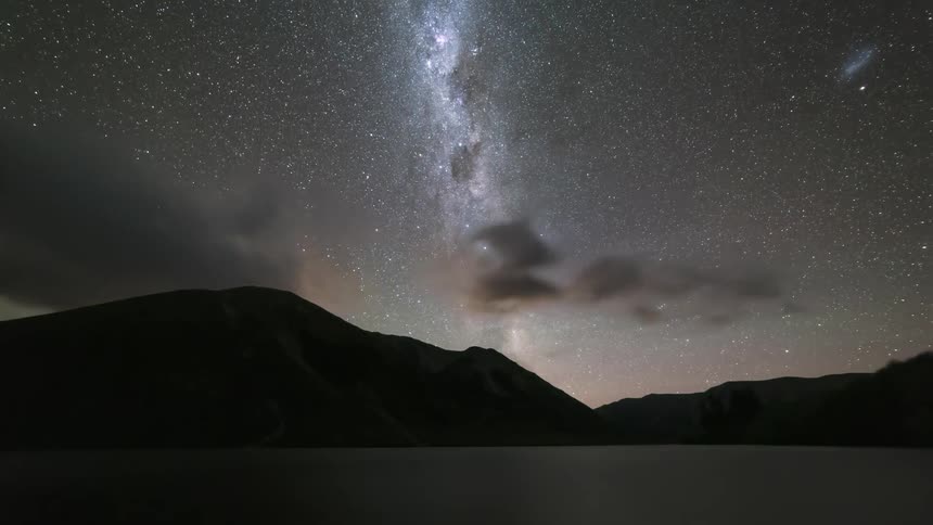 Clouds pass over the water and silhouetted mountains as darkness falls and the night sky rotates overhead