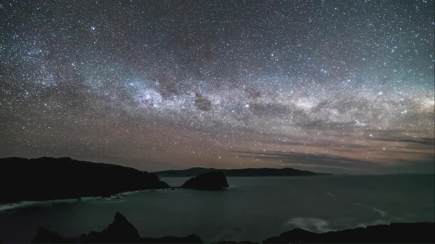 A starry sky at night on a coast and there is a green glow of the aurora borealis on the horizon