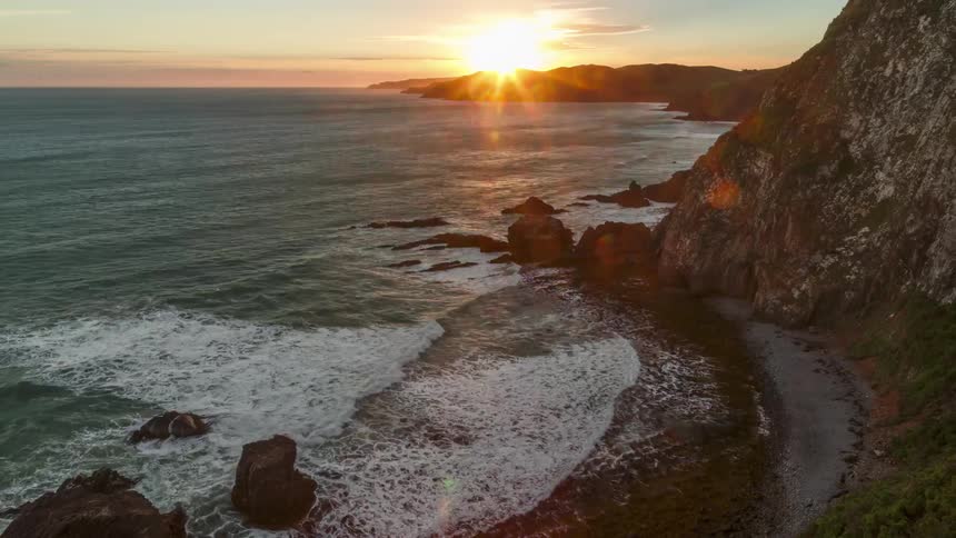 Waves foam while crashing against beach at the base of seaside cliff as the sun sets