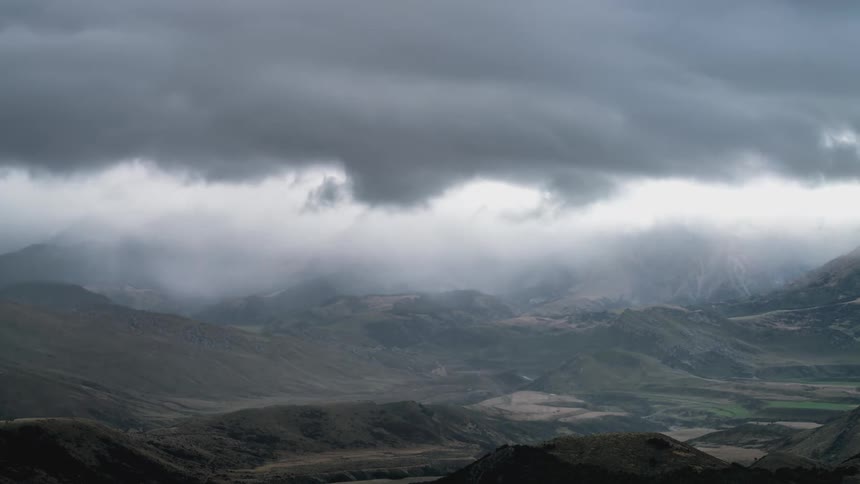 A mountainous region with many white and gray clouds overhead in the sky heading to the left