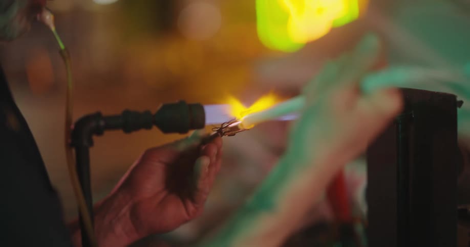 A man is melting glass using a blowtorch while blowing into the tube with his mouth