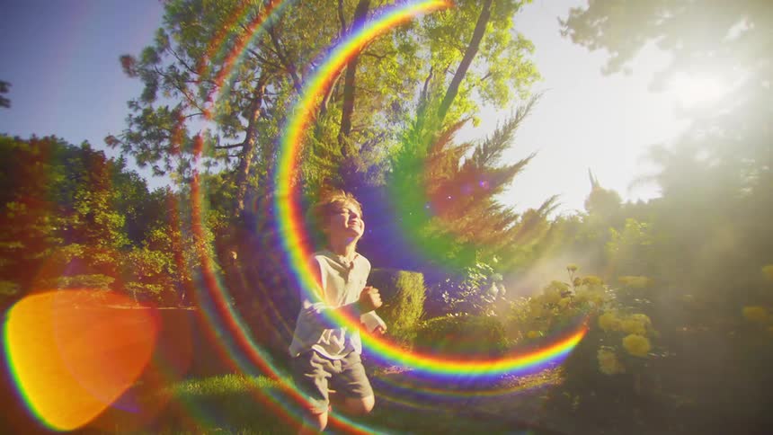A little boy is running happily on a lawn in a backyard with several trees and strong sunlight