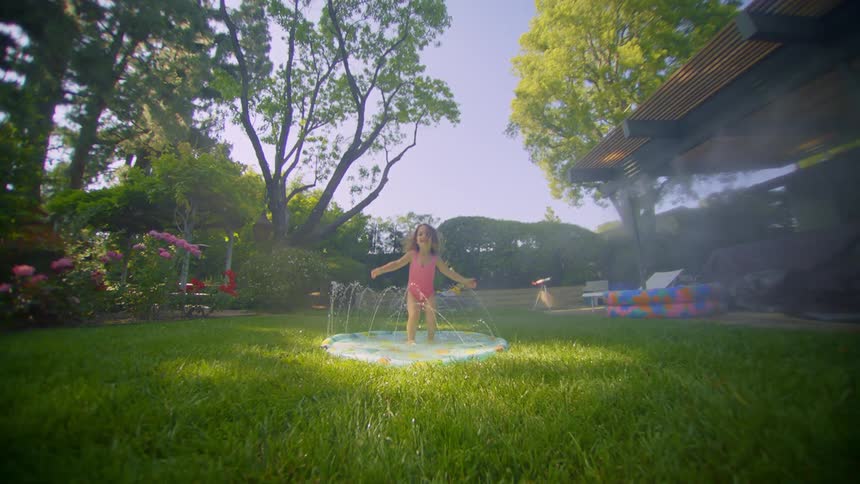 A little girl is in a little pool jumping happily in a yard with green grass