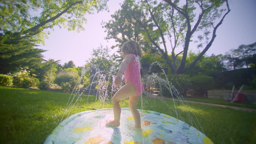 A little girl is walking in a small pool with water shooting upwards in a grassy yard