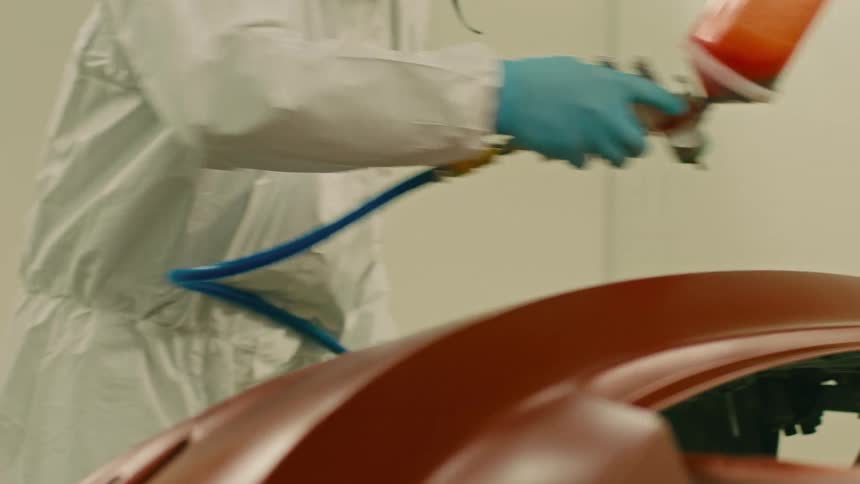 A person in a white lab coat and blue gloves is using a spray gun to apply a coating to a curved brown object