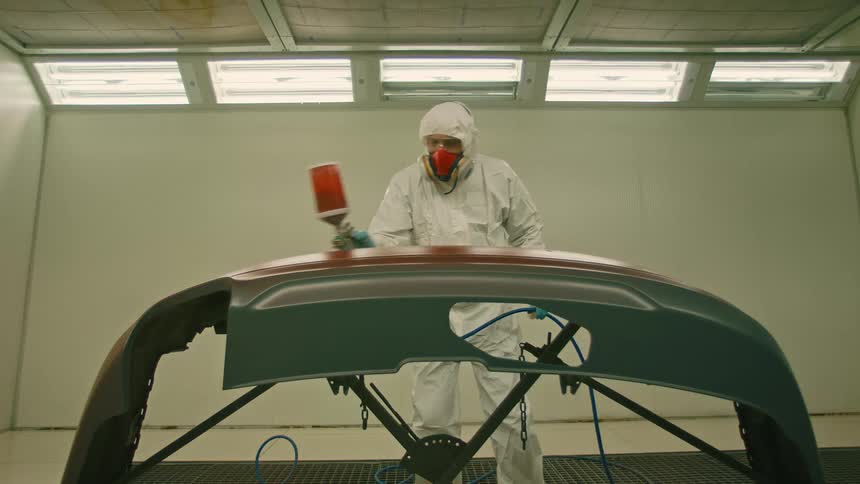 A person in protective gear is spray painting a car part in a welllit room