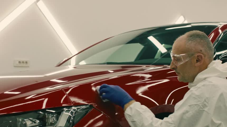 An individual in a white lab coat inspecting the finish of a red cars front panel near the headlight