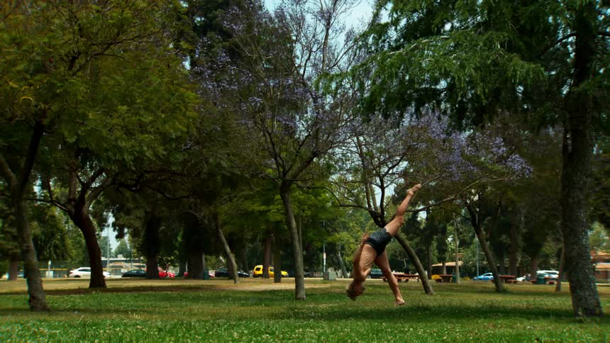 A shirtless man is in a grass park with trees doing ballet steps and doing a forward somersault