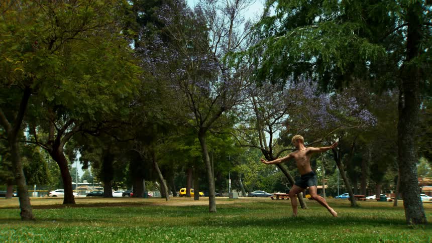 A blond man is shirtless and doing ballet steps in a park with grass and several trees