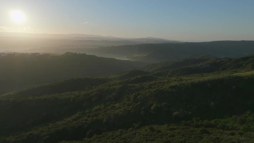 An aerial view of a region with hills covered in green trees and fog on the horizon and a bright sun in the sky
