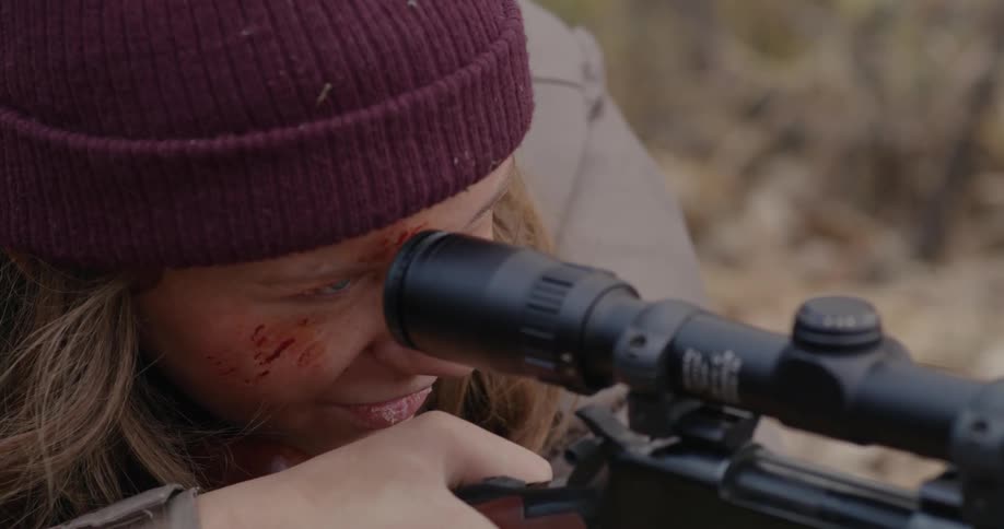 A woman with a scraped up face looks through a rifle scope and fires