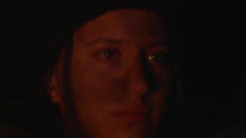 A young womans face that is dirty and she is looking forward where there is a fire torch illuminating the dark environment