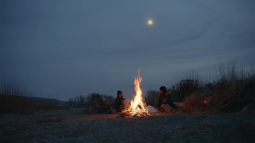 Two people sitting around a bonfire with tall orange flames on an evening