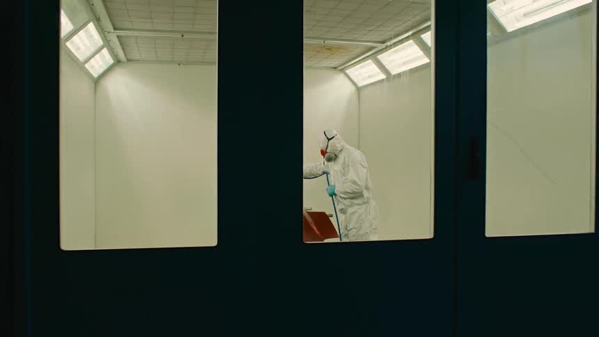 Automotive painter sprays a fender in a garage while wearing protective gear