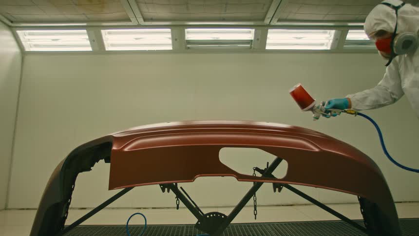 A person in white protective clothing using a paint hose to paint a car bumper brown