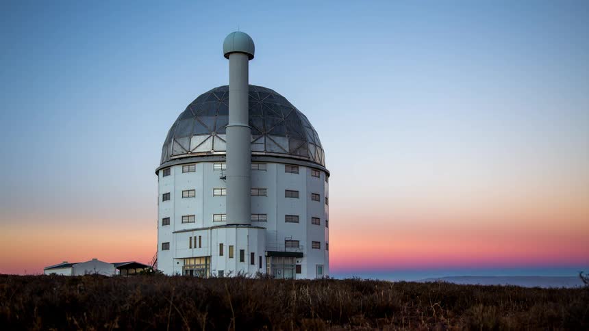 Awhite observatory is in a place pointed towards the sky that is starry and then it dawns and the sky turns orange and pink