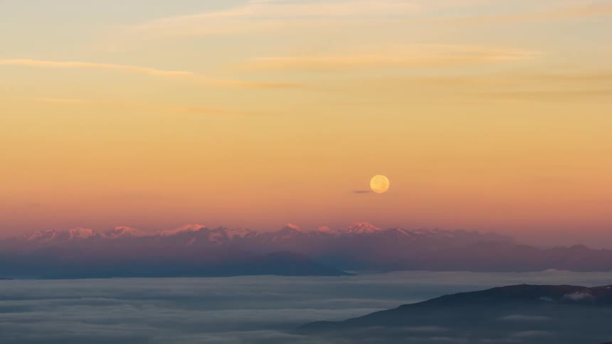 Serene landscape at dawn with layers of clouds over hills and a sky that dawns with the setting moon